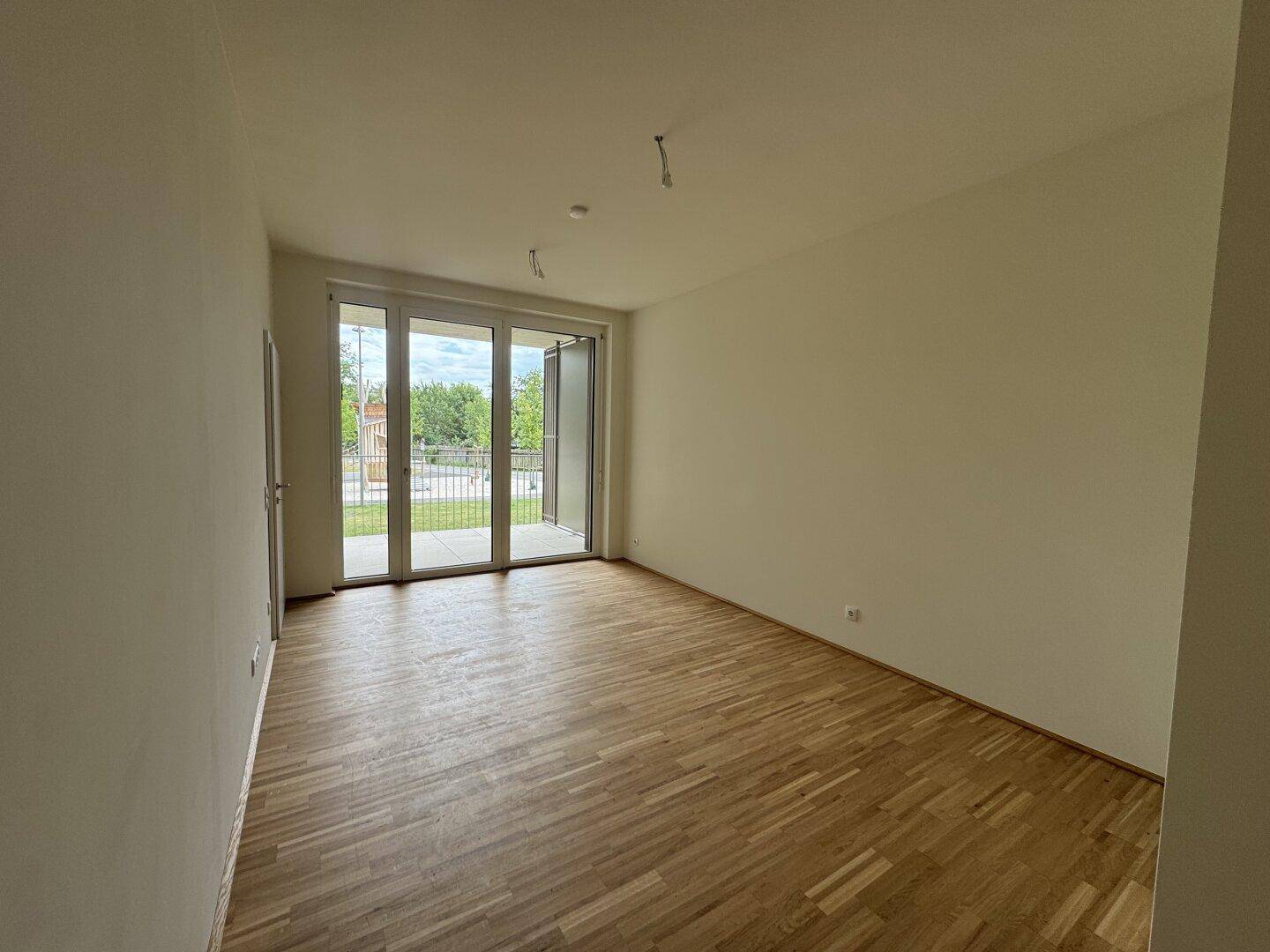 Heller, leerer Wohnraum mit Parkettboden und großen Fenstern zum Balkon.