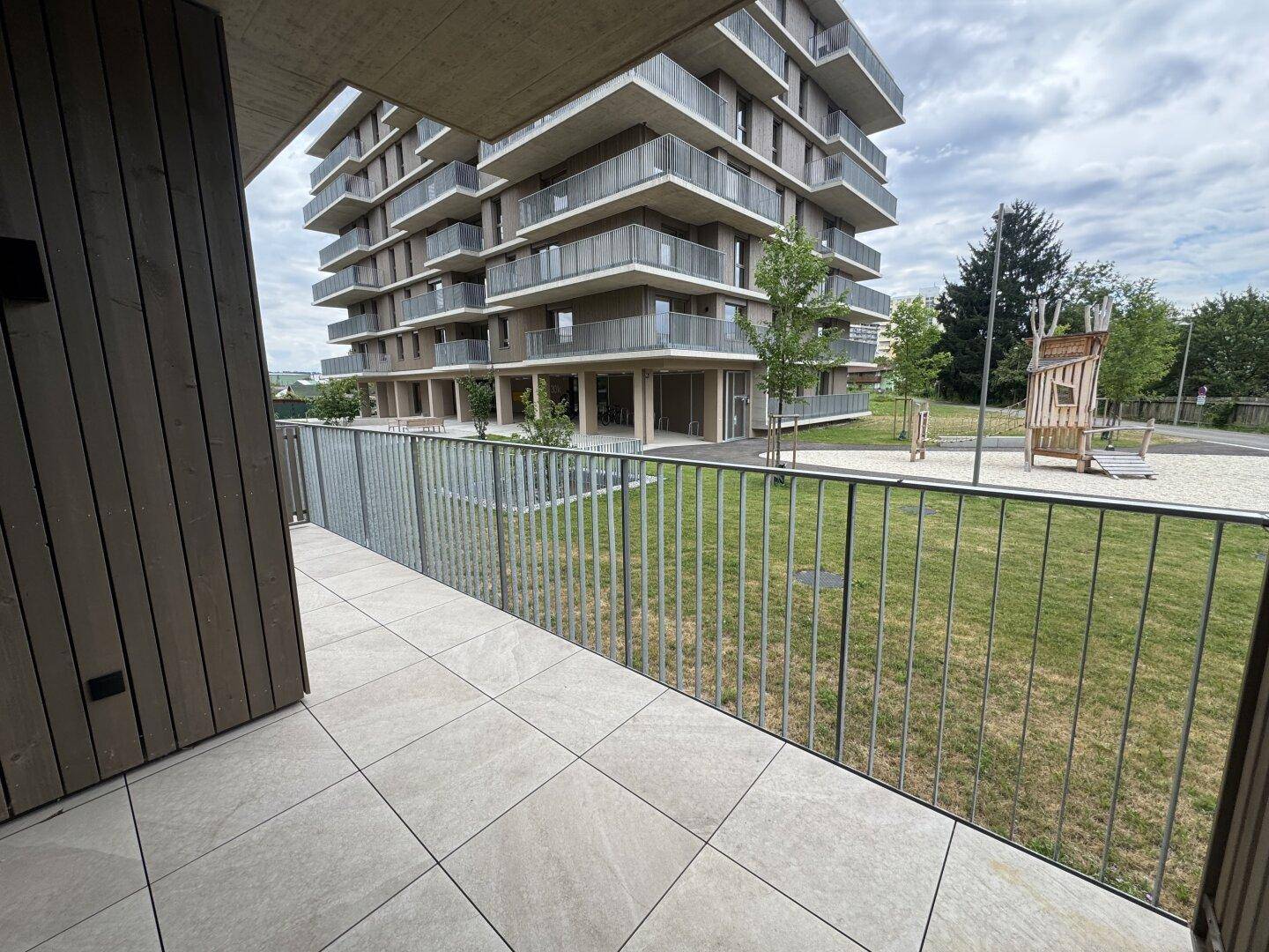 Balkon mit Fliesenboden und Blick auf das moderne Gebäude und den angrenzenden Spielplatz.