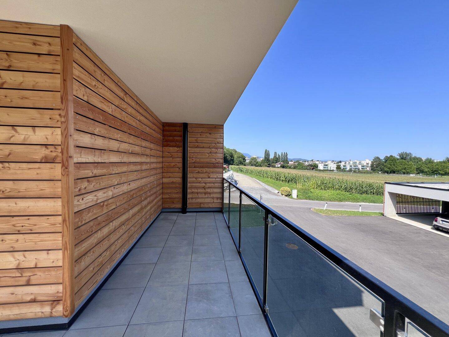 Langer Balkon mit Holzverkleidung, Glasgeländer und weitem Blick auf die Felder und Landschaft.