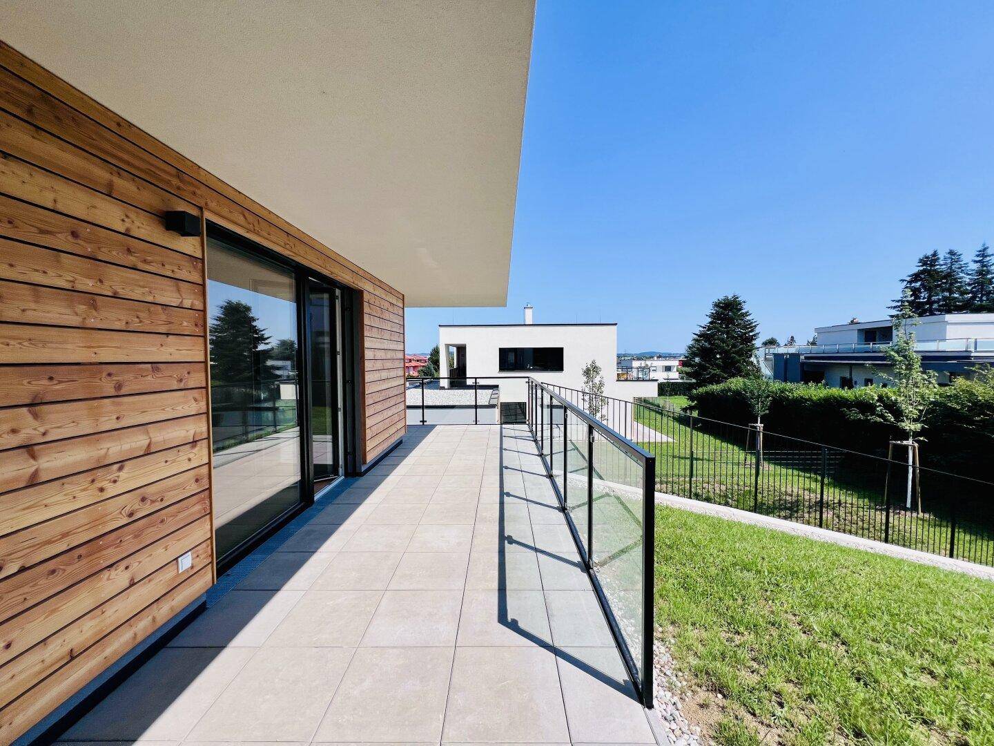 Helle Terrasse mit Fliesenboden, Glasgeländer und direktem Zugang zum grünen Gartenbereich.