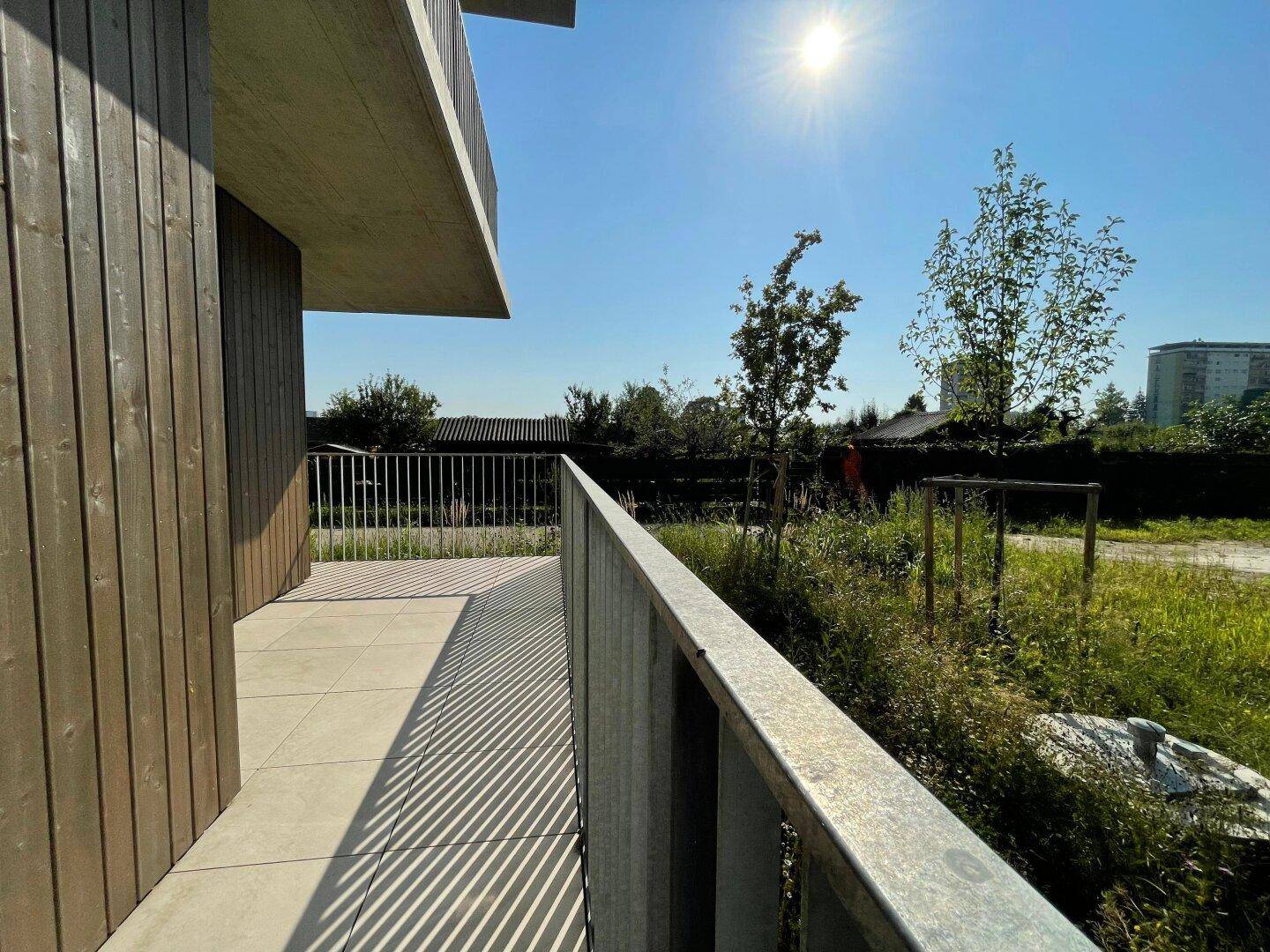 Heller Terrassenbereich mit Holzfassade und viel Sonnenschein, umgeben von Natur und Grünflächen.
