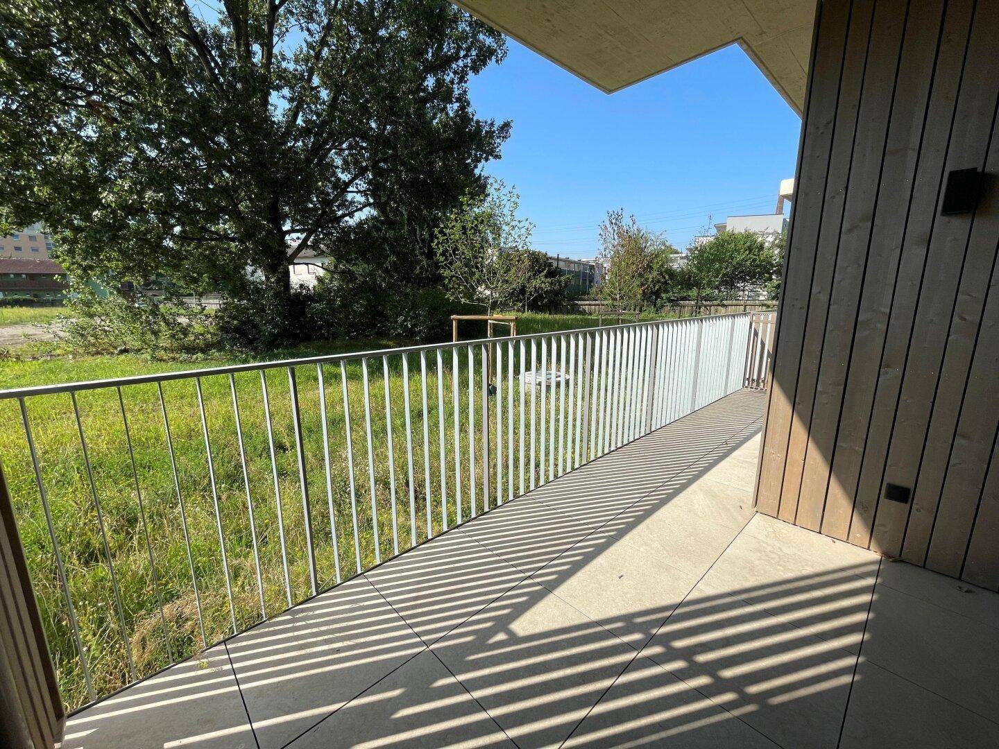 Sonniger Balkon mit Fliesenboden und Metallgeländer, umgeben von viel Grün und Bäumen.