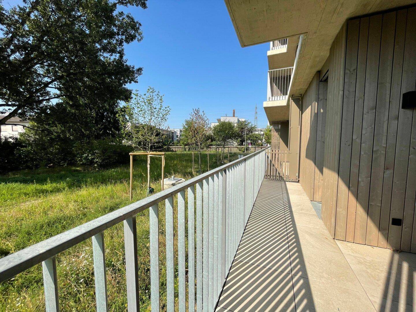 Langer Balkon mit Holzfassade und Blick auf die grüne Umgebung und weitere Gebäude.
