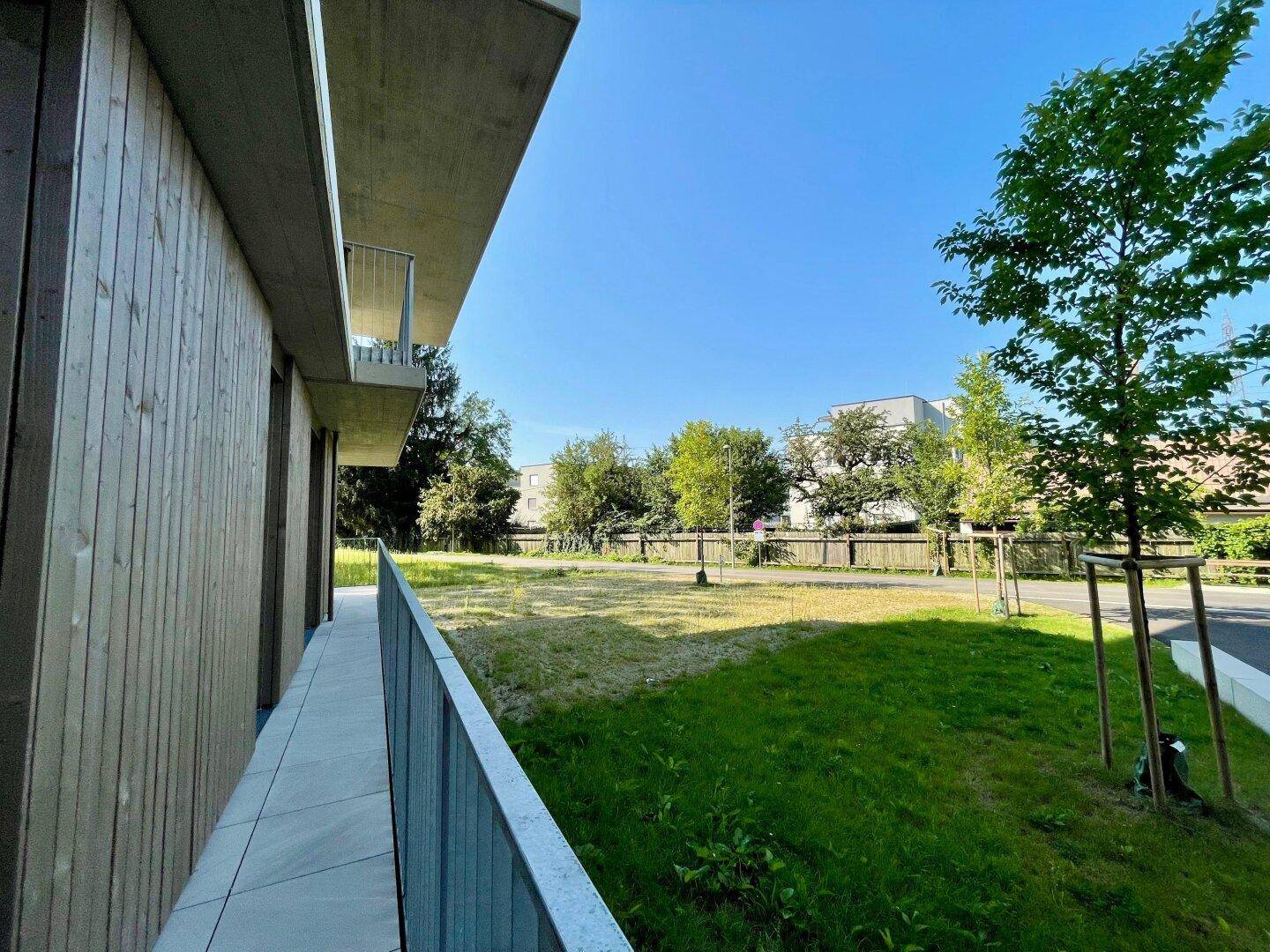 Balkon eines modernen Gebäudes mit seitlichem Blick auf eine gepflegte Rasenfläche und junge Bäume.