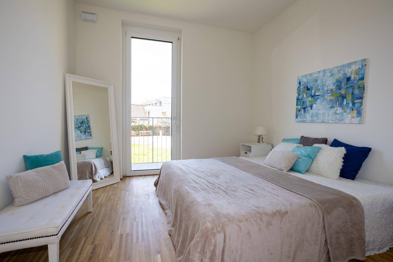 Helles Schlafzimmer mit Holzparkett, großem Bett und Zugang zu einem Balkon.