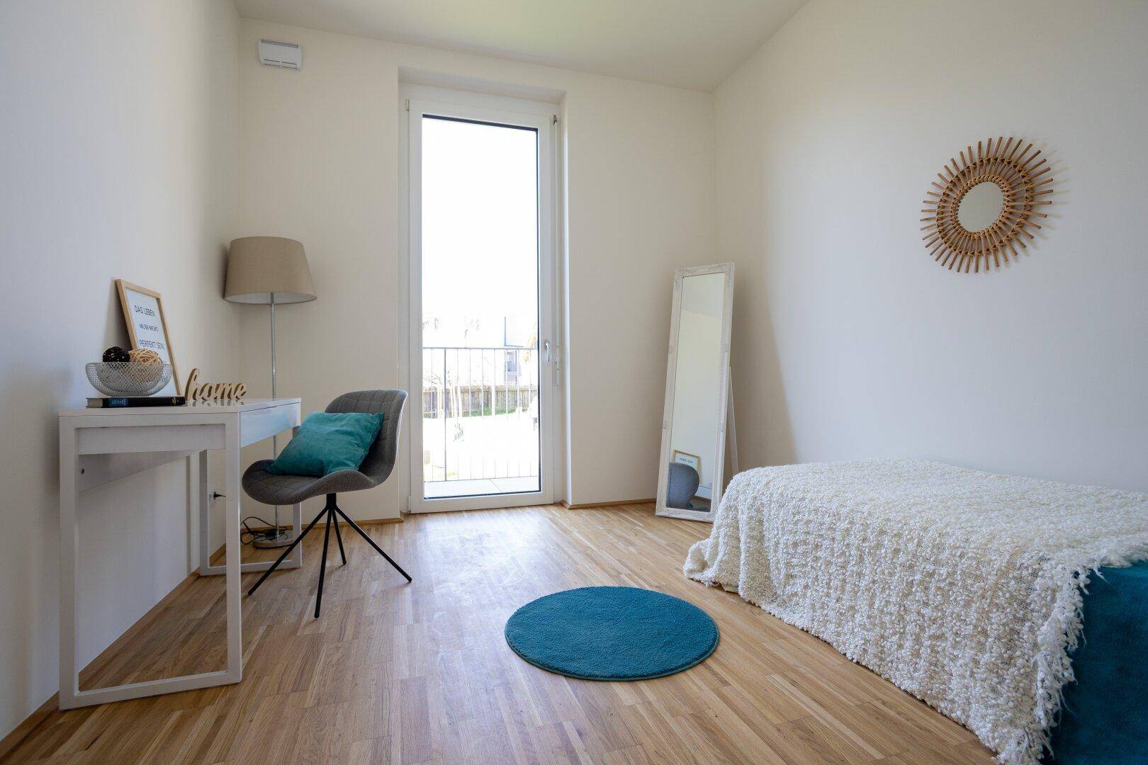Freundliches Zimmer mit Holzparkett, Schreibtisch und Zugang zu einem Balkon.