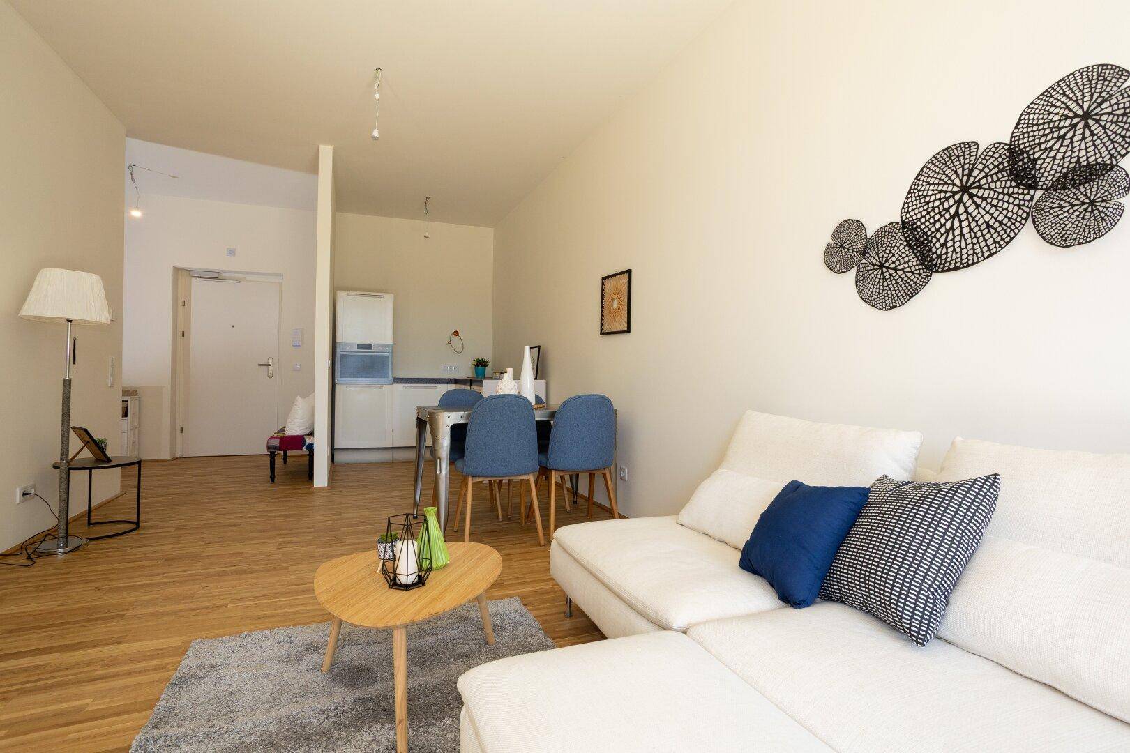 Offener Wohn-Essbereich mit Holzboden, weißem Sofa und integrierter Küchenzeile im Hintergrund.