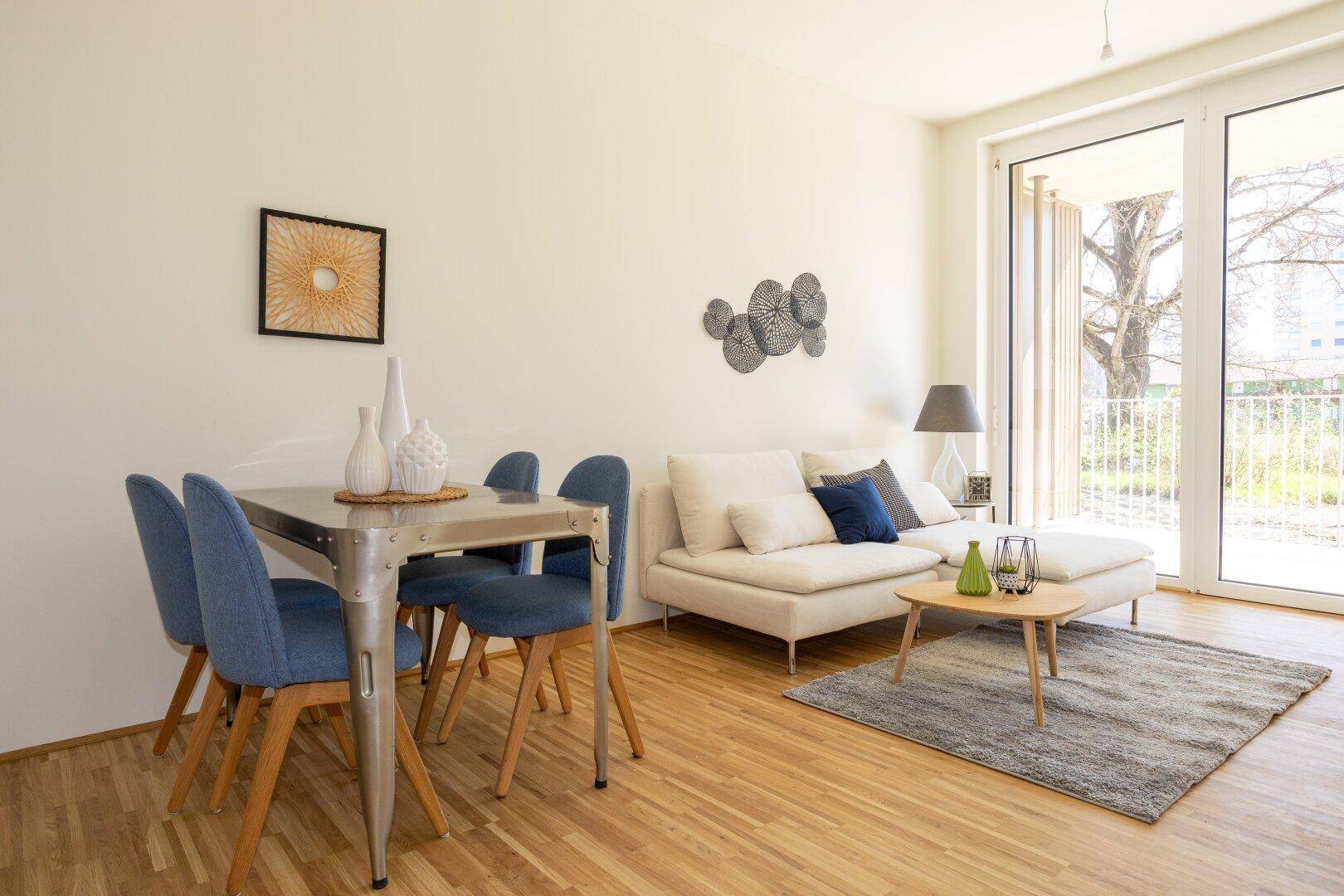 Lichtdurchfluteter Wohn-Essbereich mit Holzboden, modernem Sofa und Zugang zum Balkon.