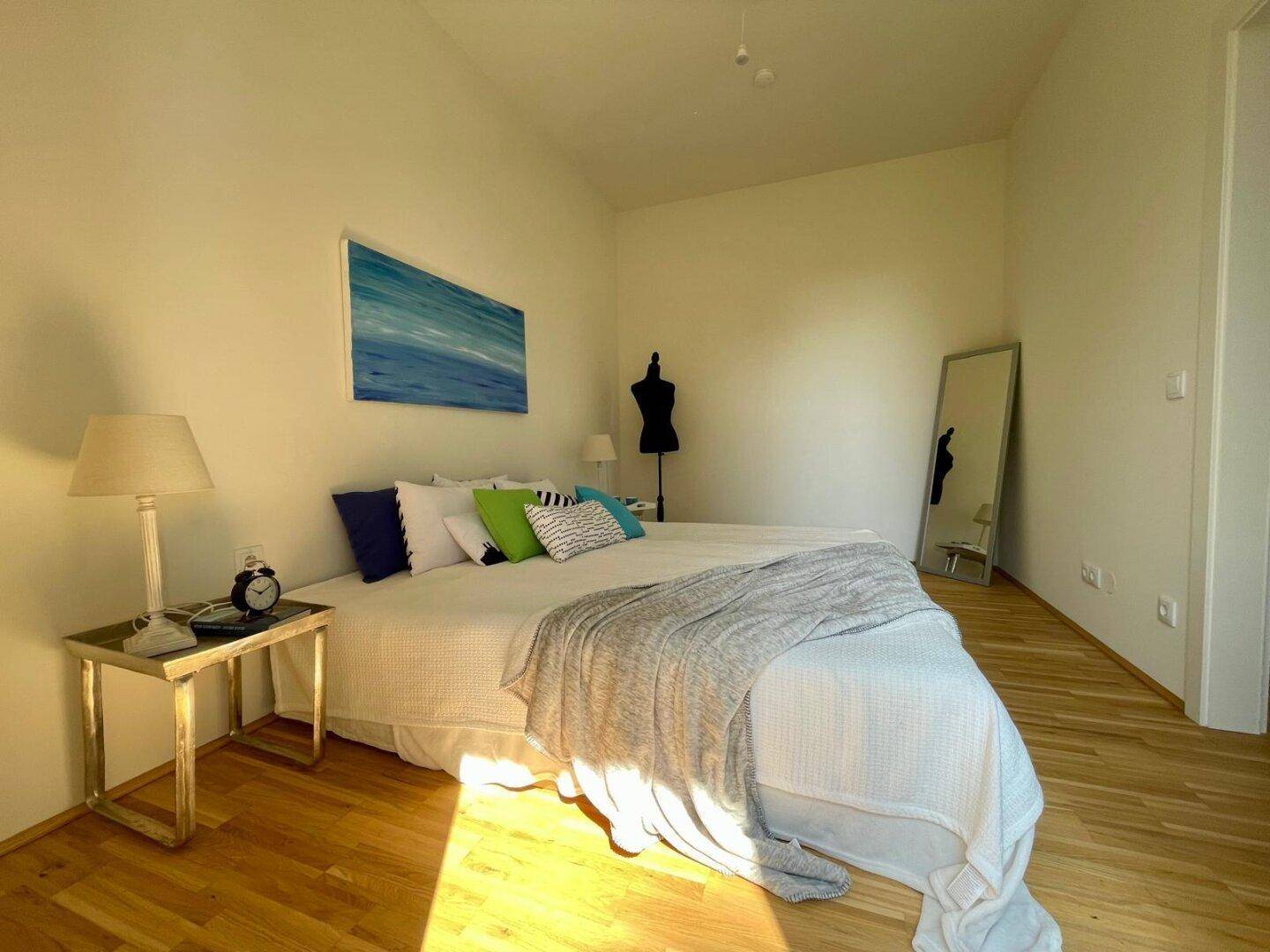 Helles Schlafzimmer mit Holzboden, großem Bett und farbigen Akzenten, sonnendurchflutet.