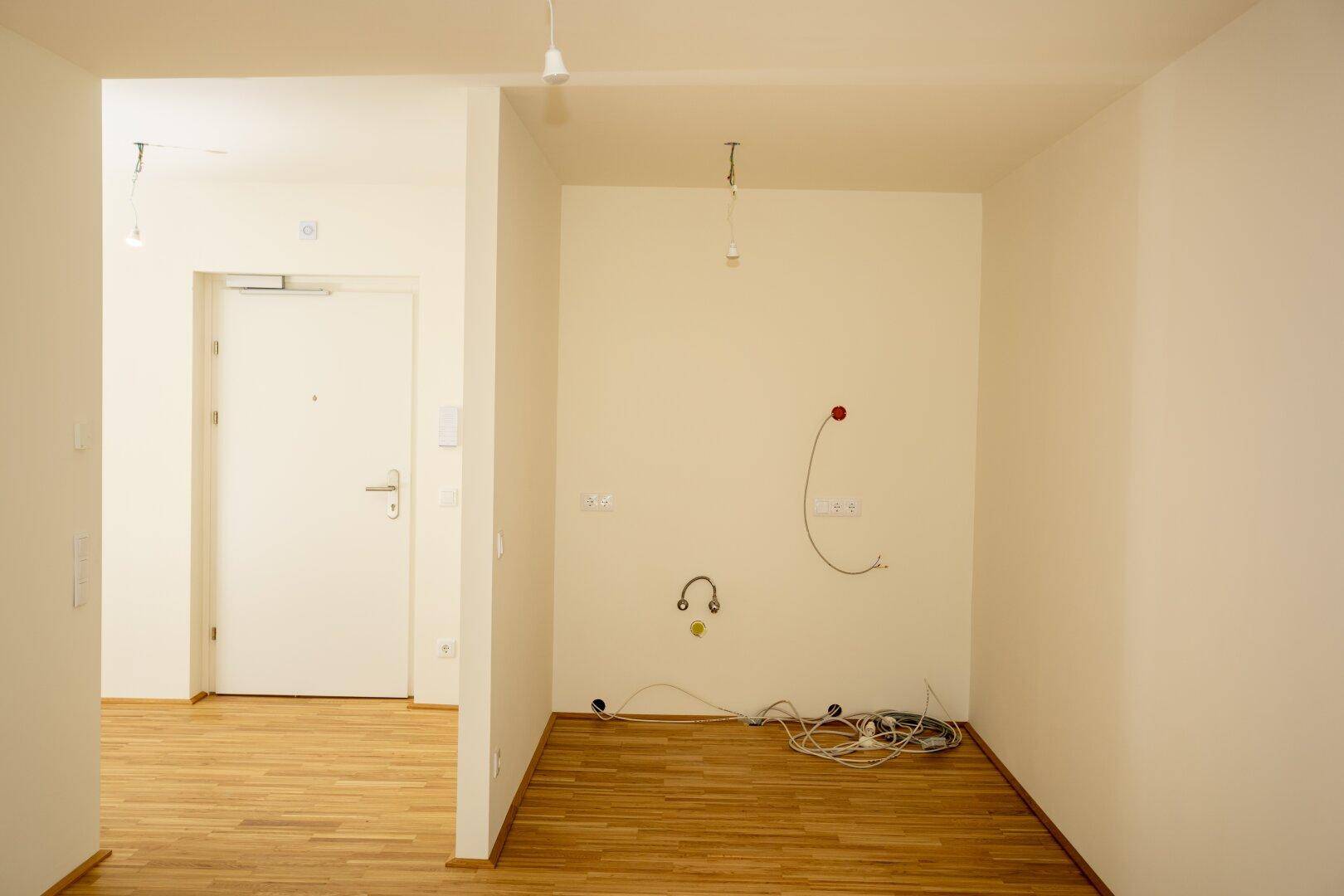 Leerer Küchenbereich mit Holzboden und vorbereiteten Anschlüssen für die Installation.