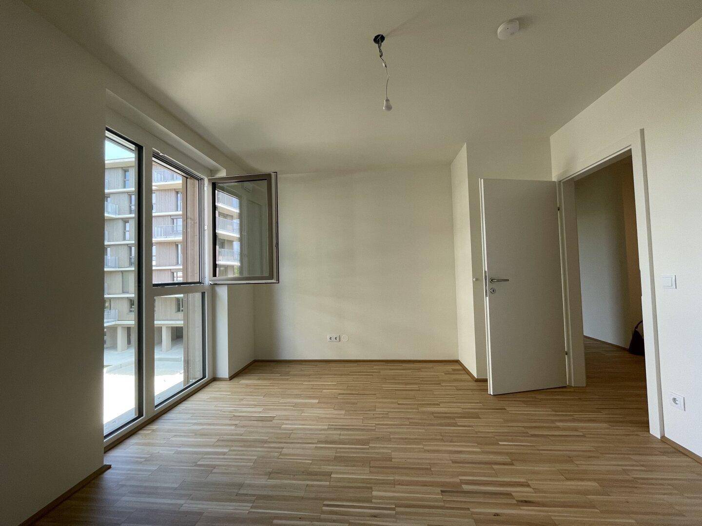 Helles Zimmer mit Parkettboden und großen Fenstern, die viel Tageslicht hereinlassen.