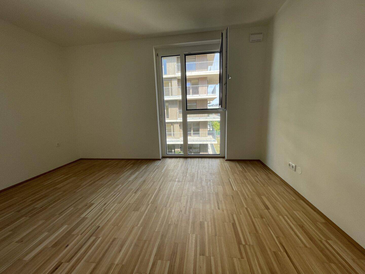 Unmöbliertes Zimmer mit Parkettboden und einer großen Fensterfront mit Blick nach draußen.