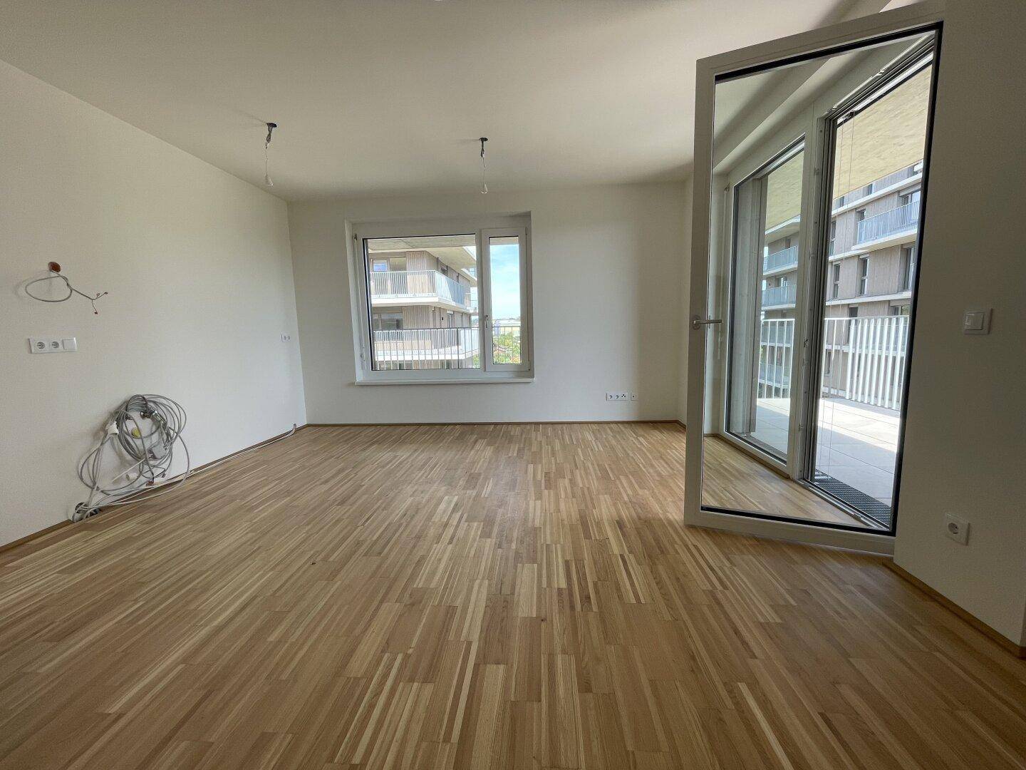 Leeres Zimmer mit Parkettboden, großen Fenstern und Zugang zu einem Balkon.