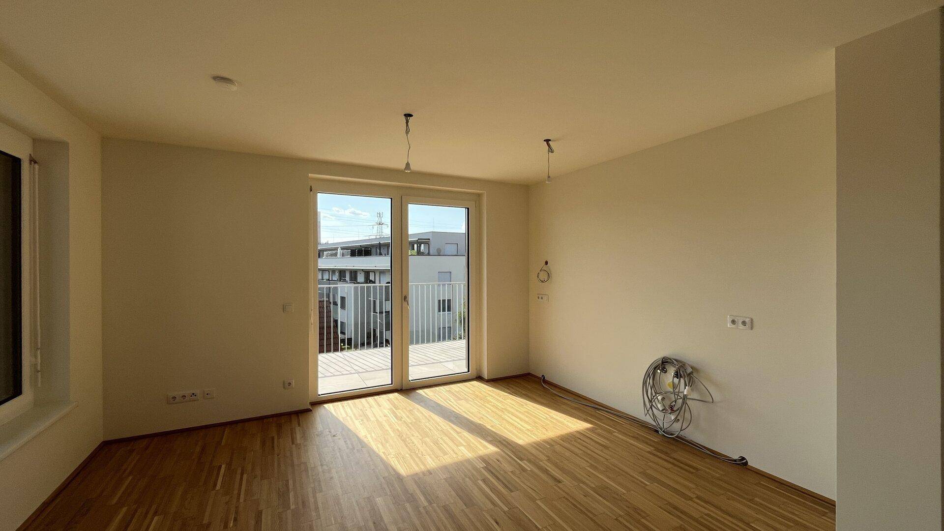 Sonniger Wohnraum mit hellem Parkettboden, weißen Wänden und direktem Zugang zum Balkon.