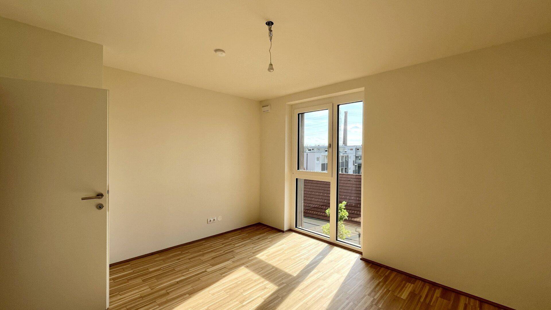Helles Schlafzimmer mit Parkettboden und großem Fenster, das Zugang zu einem kleinen Außenbereich bietet.
