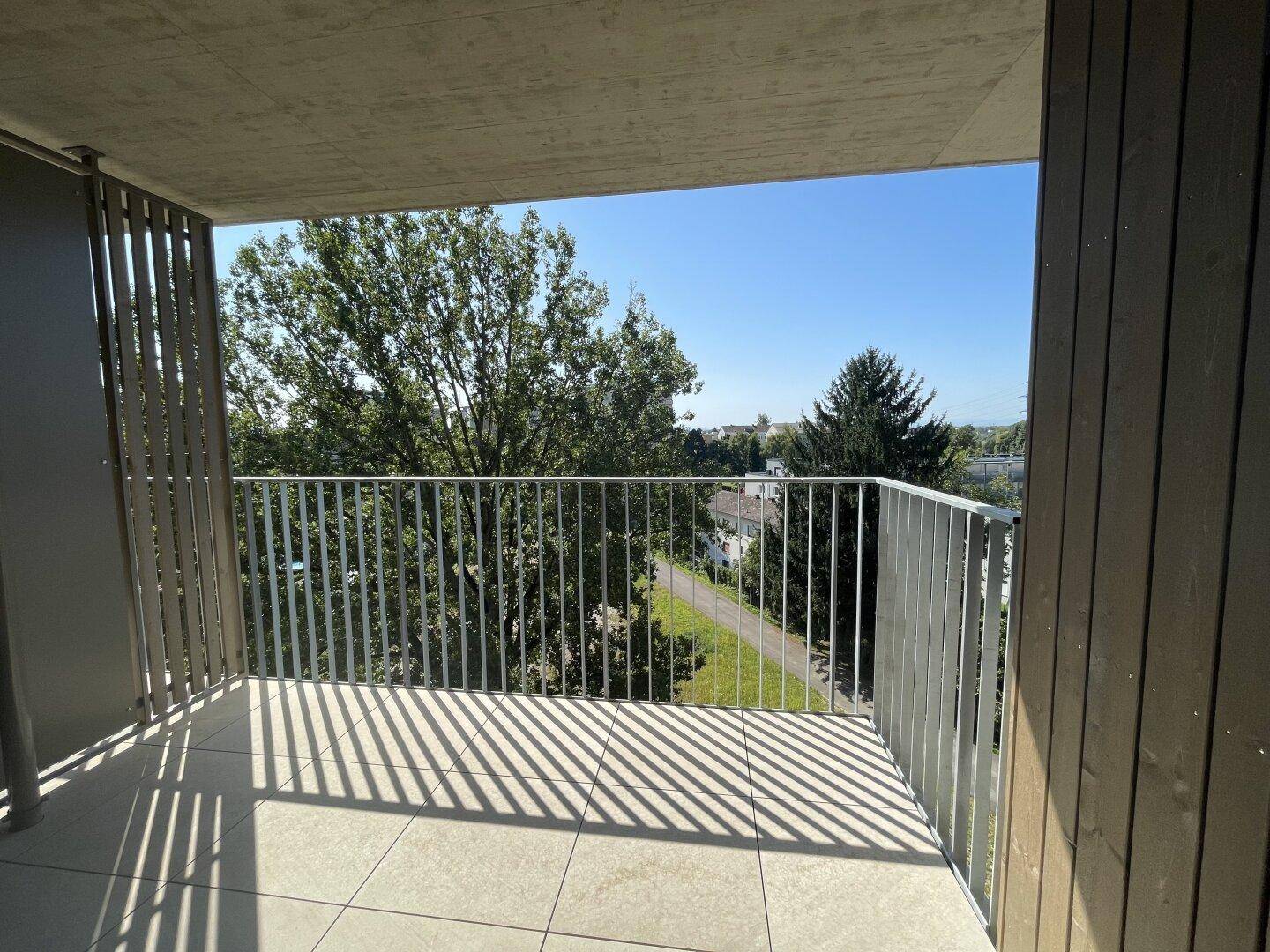 Sonniger Balkon mit Fliesenboden und Blick auf grüne Bäume und einen Spazierweg.