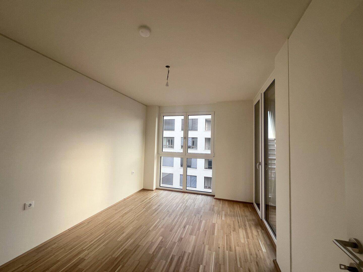 Leeres Schlafzimmer mit Parkettboden und großem Fenster, das viel Tageslicht hereinlässt.