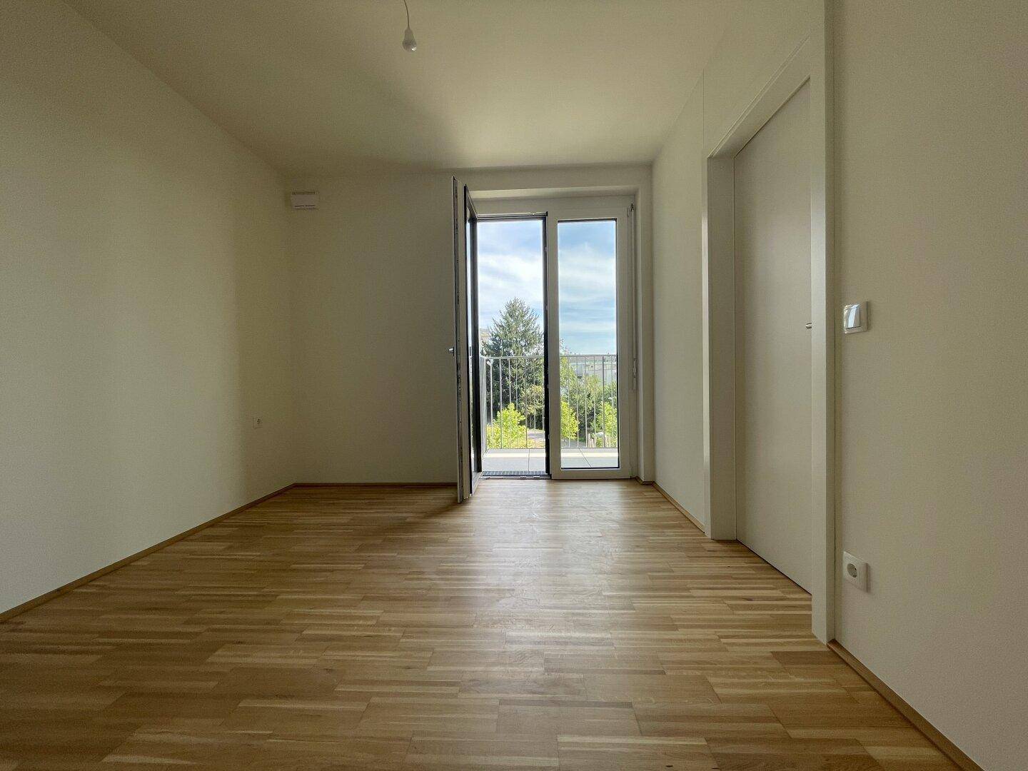 Heller, leerer Raum mit Holzfußboden und Zugang zu einem kleinen Balkon mit Grünblick.