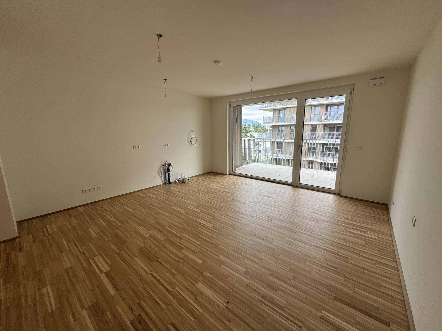 Helles Wohnzimmer mit Parkettboden und Zugang zu einem großen Balkon in einem Neubau.