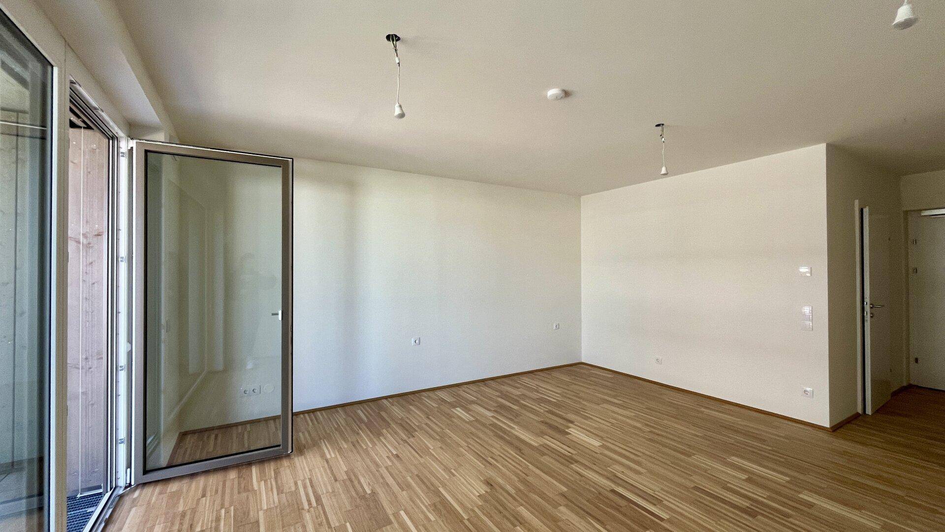 Geräumiges Wohnzimmer mit Holzboden und viel Tageslicht durch die Balkontür in einem Neubau.