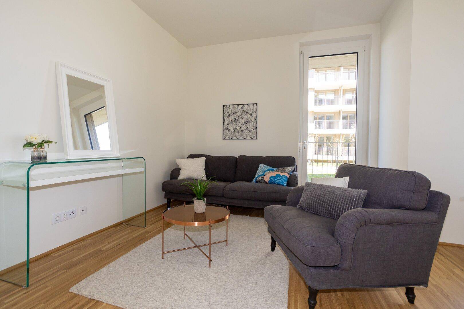 Gemütliches Wohnzimmer mit dunklem Sofa, Sessel und Zugang zu einem Außenbereich.