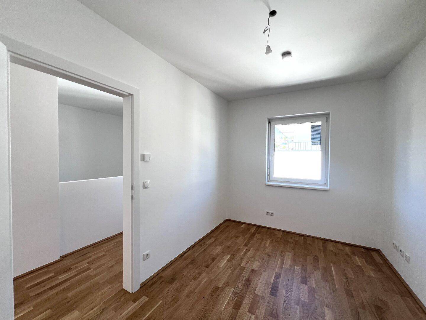 Helles Schlafzimmer mit Parkettboden und einem Fenster, das Tageslicht hereinlässt.