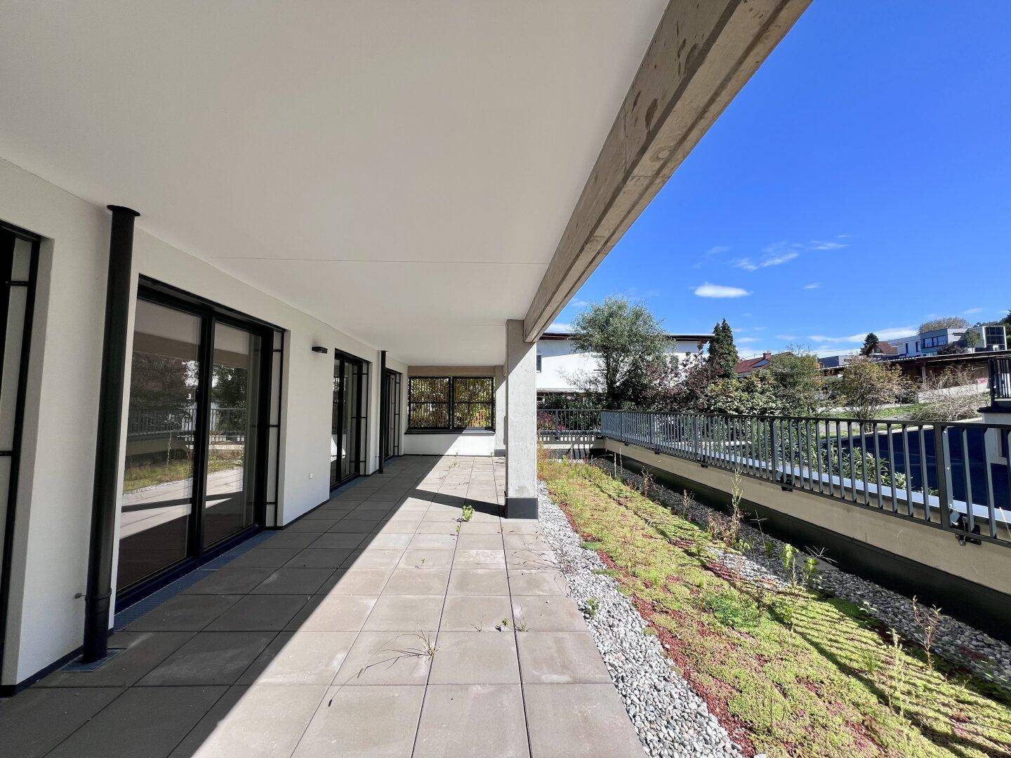 Lange, geflieste Terrasse mit Grünfläche und Blick auf die Umgebung unter blauem Himmel.