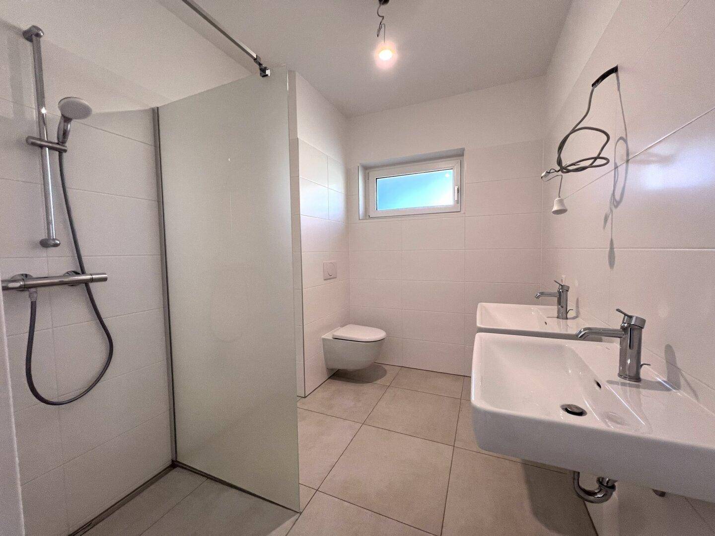 Modernes Badezimmer mit Doppelwaschbecken, Dusche, WC und einem Fenster für Tageslicht.