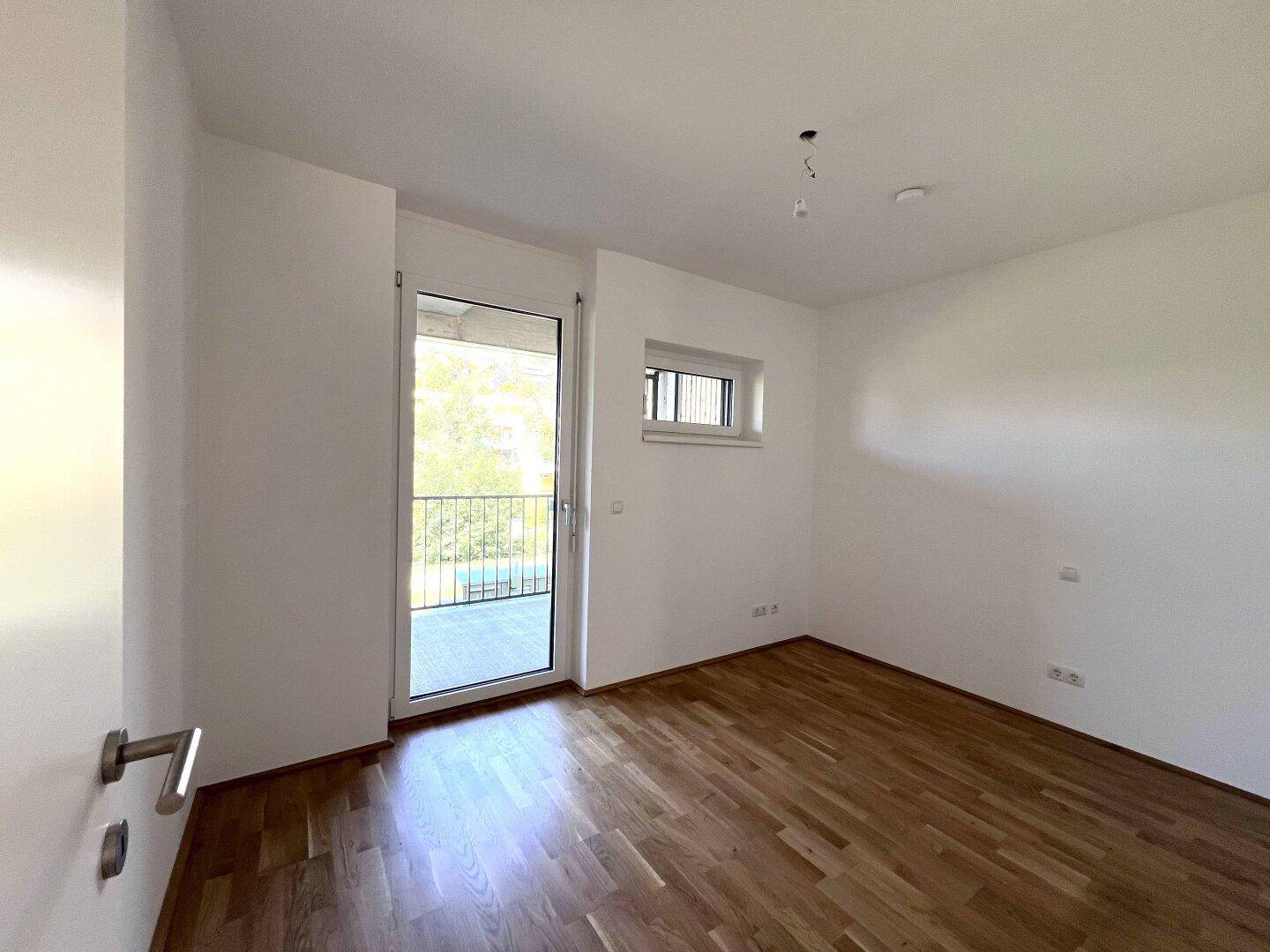 Helles Schlafzimmer mit Holzboden, einem Fenster und Zugang zu einem Außenbereich.