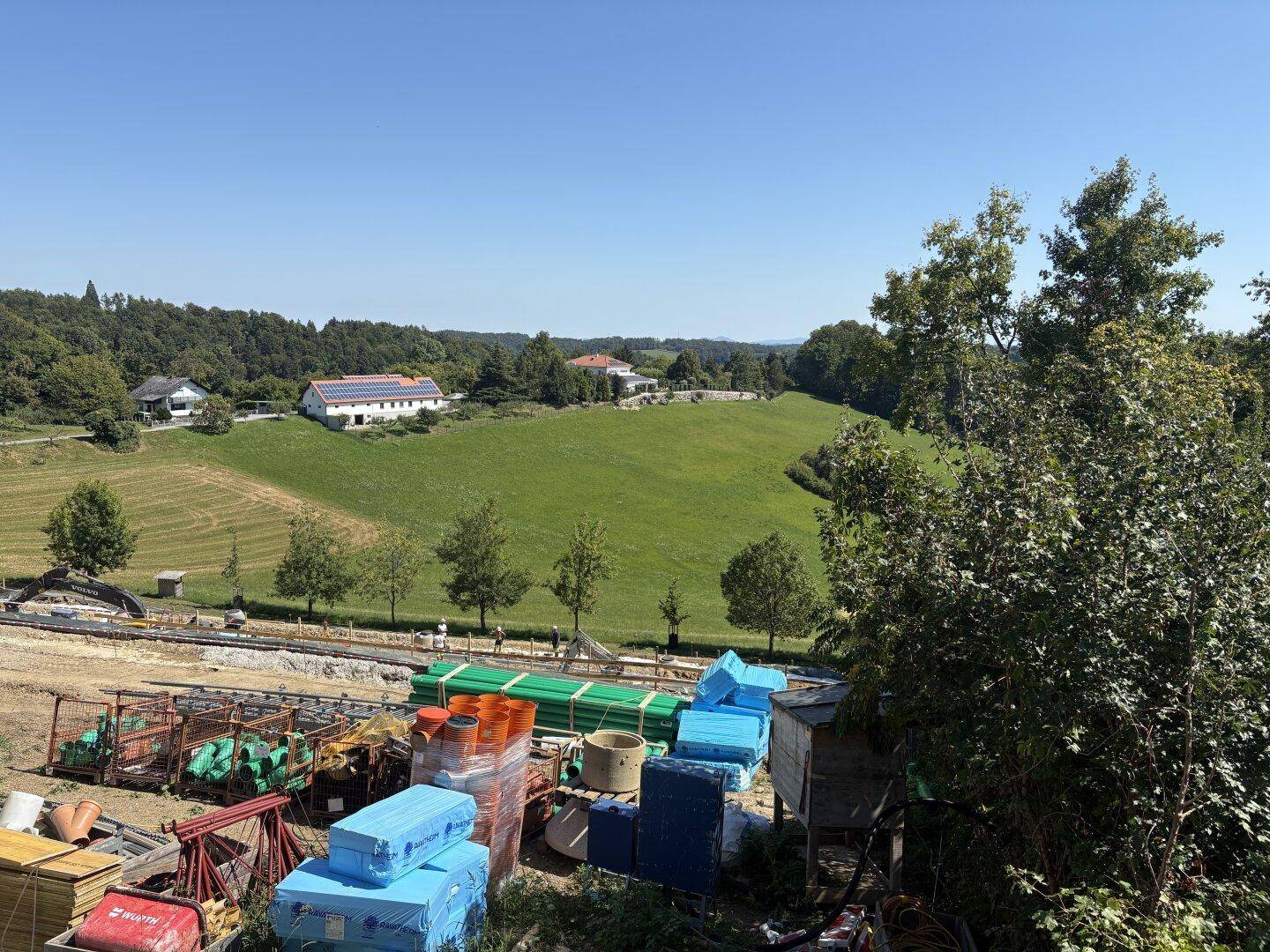 Baustelle mit Materialien im Vordergrund und idyllischer grüner Landschaft im Hintergrund.
