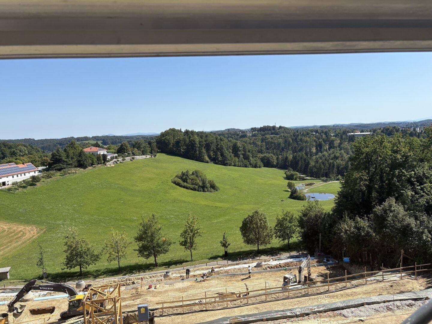 Blick auf eine große Baustelle mit Erdarbeiten und eine weite grüne Landschaft mit Teich.