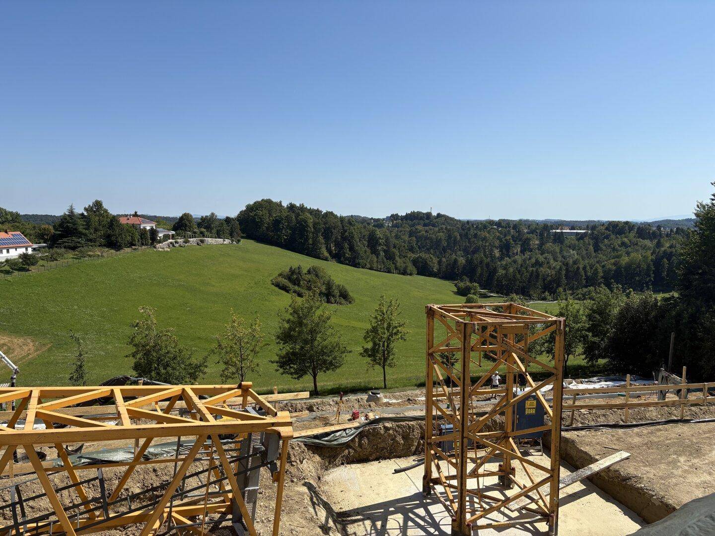 Baustelle mit Kranstruktur im Vordergrund und weitläufiger grüner Landschaft im Hintergrund.