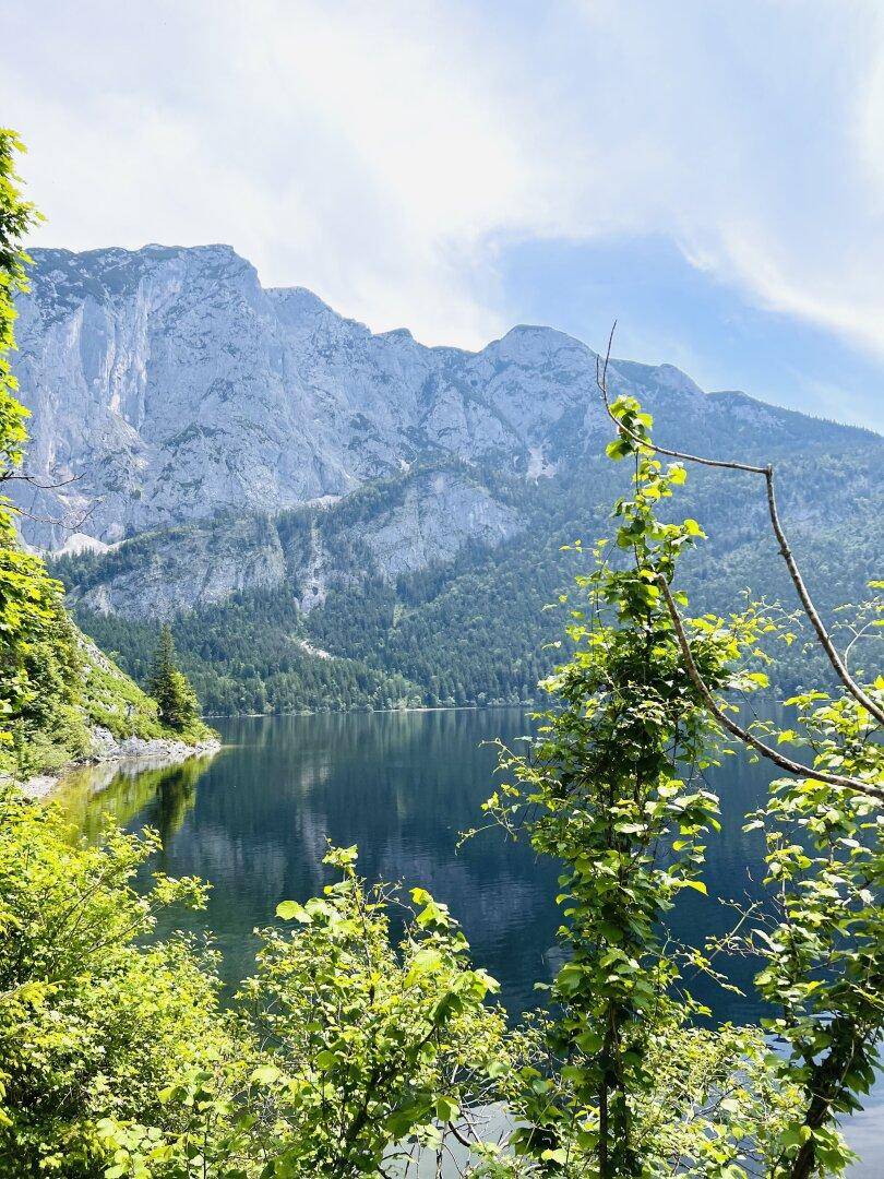 Ruhiger Bergsee umgeben von üppigem Grün und imposanten Felswänden unter einem klaren blauen Himmel.