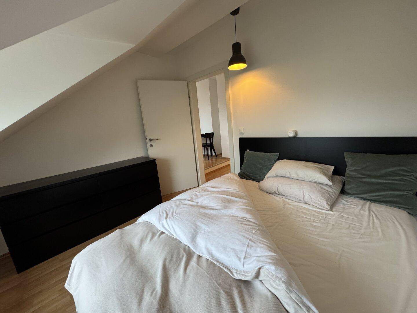 Schlafzimmer mit Holzboden und dunkler Kommode, verbunden mit dem angrenzenden Wohnbereich.