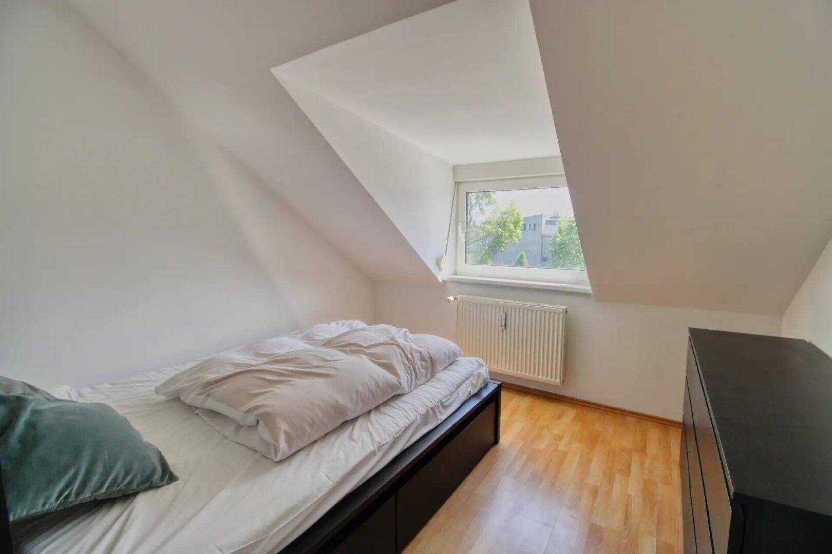 Helles Schlafzimmer im Dachgeschoss mit Holzboden und einem Fenster mit Blick ins Grüne.