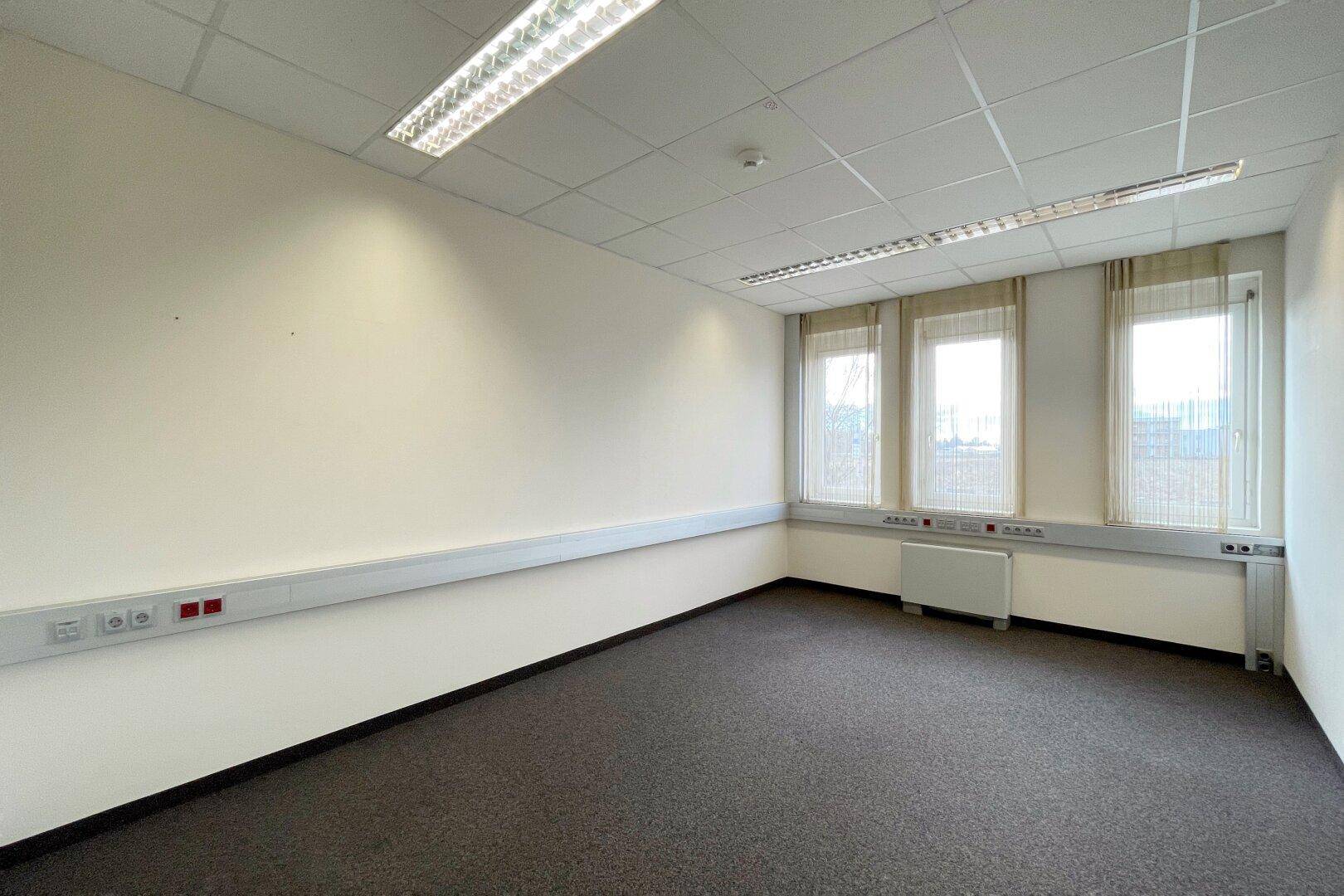 Funktionales Büro mit Teppichboden, Kabelkanälen und Fenstern, die für ausreichend Helligkeit sorgen.