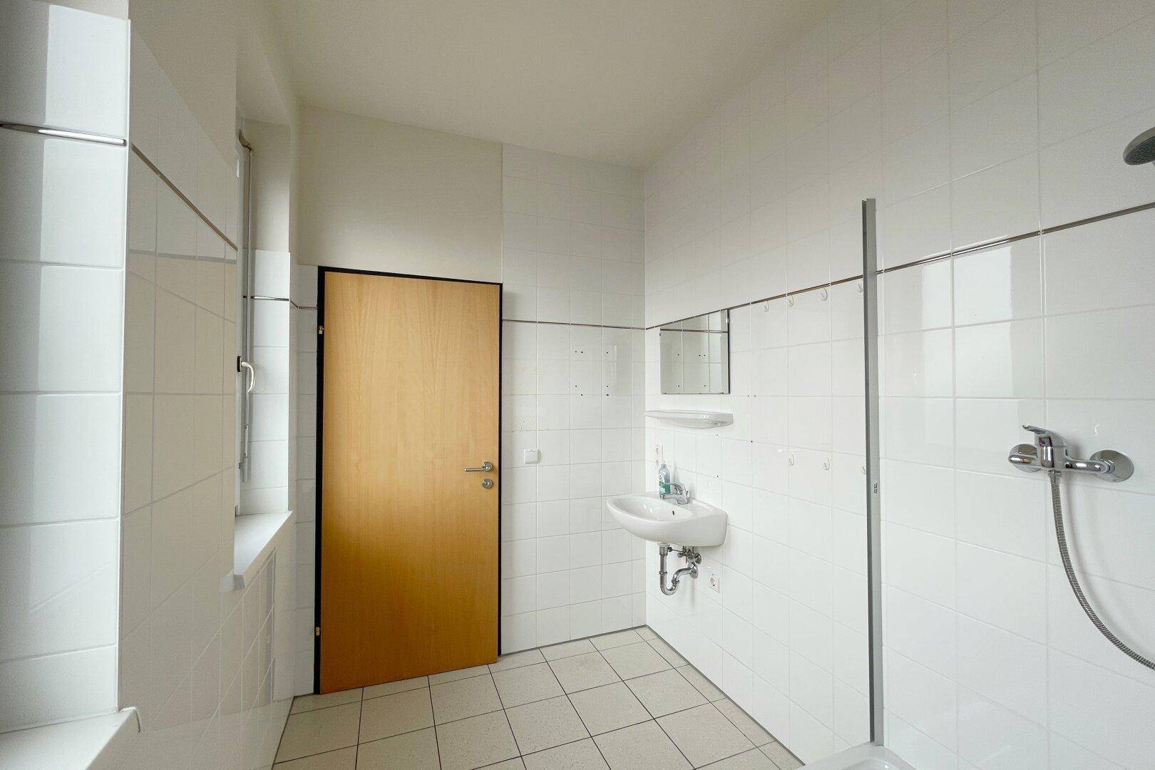 Funktionales Badezimmer mit Duschbereich, Waschbecken und Fenster, komplett gefliest in Weiß.