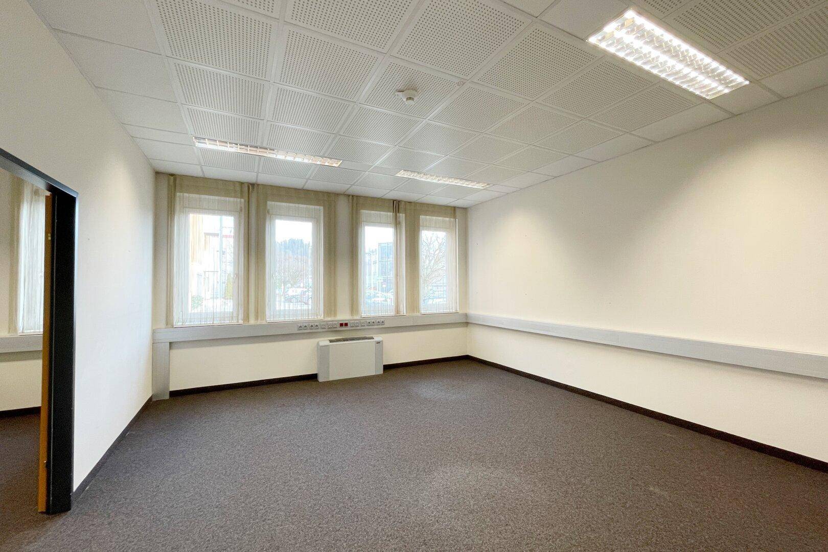 Helles Büro mit Teppichboden, abgehängter Decke und mehreren Fenstern für eine angenehme Arbeitsatmosphäre.