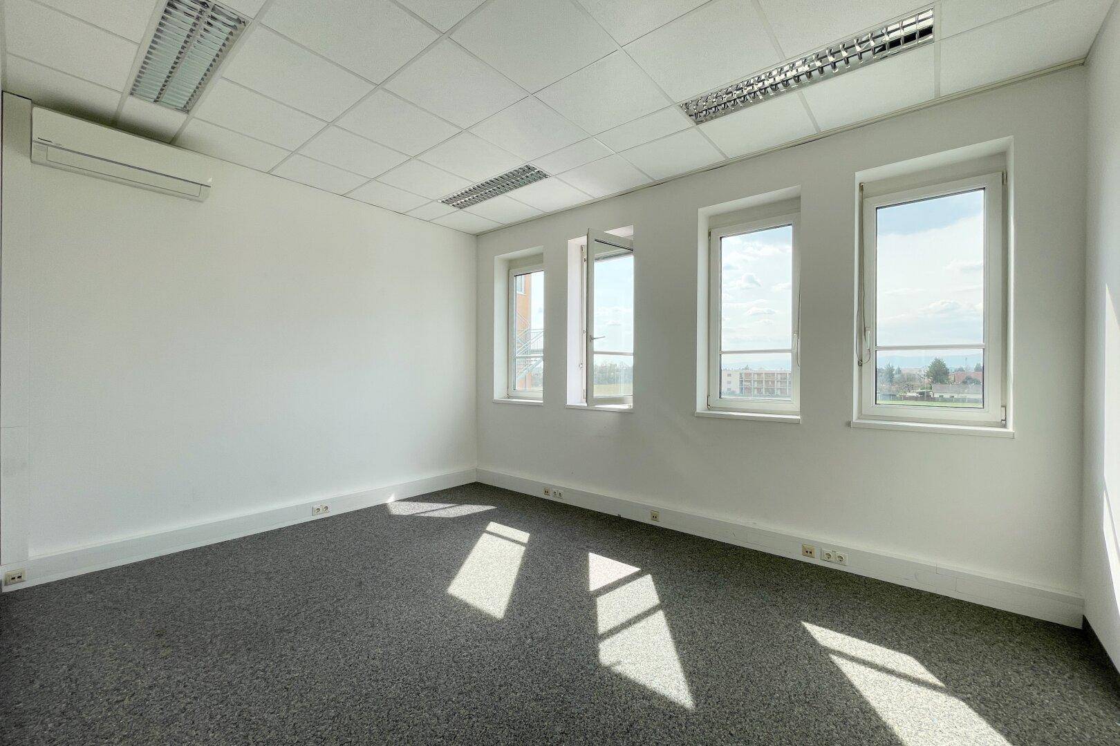 Helles und sauberes Büro mit Teppichboden, weißen Wänden und drei Fenstern, die viel Licht spenden.
