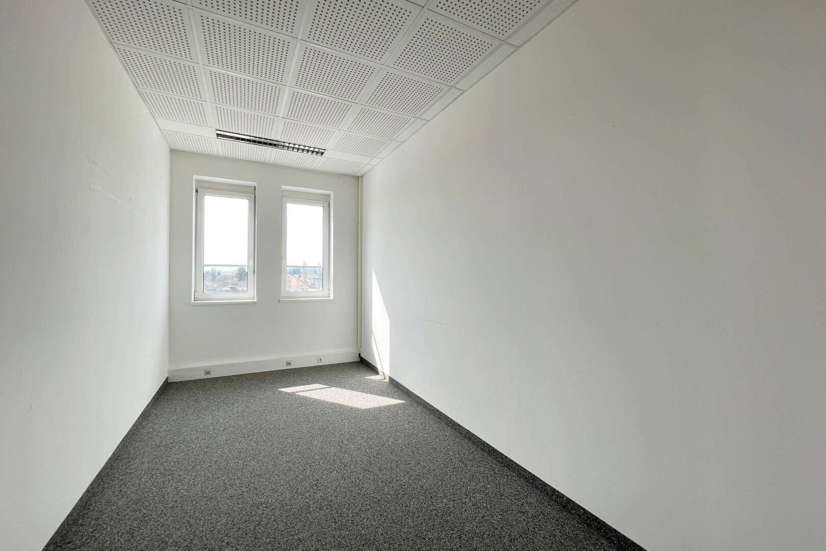 Kleineres, helles Büro mit Teppichboden, weißen Wänden und zwei Fenstern, die Tageslicht hereinlassen.