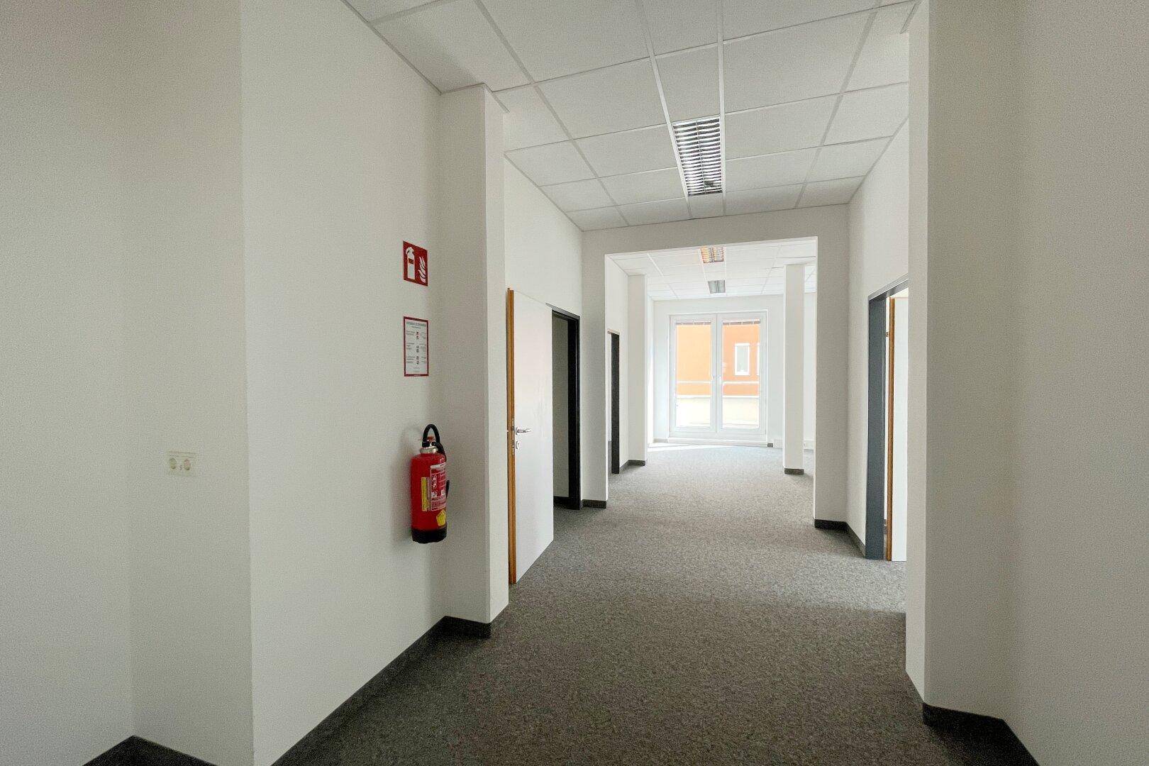 Langer, heller Flur mit Teppichboden, weißen Wänden und Deckenplatten in einem Bürogebäude.