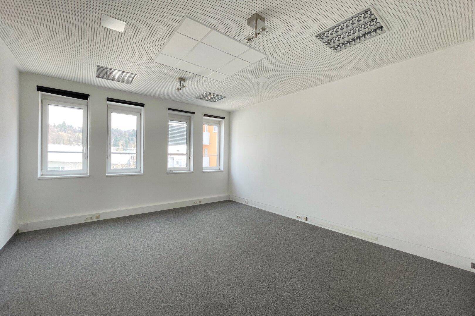 Leeres Büro mit Teppichboden, weißen Wänden, abgehängter Decke und vier Fenstern mit Rollos.