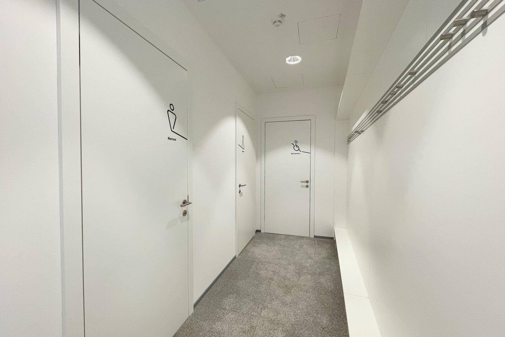 Heller Flur mit Teppichboden und Türen zu barrierefreien Toiletten.