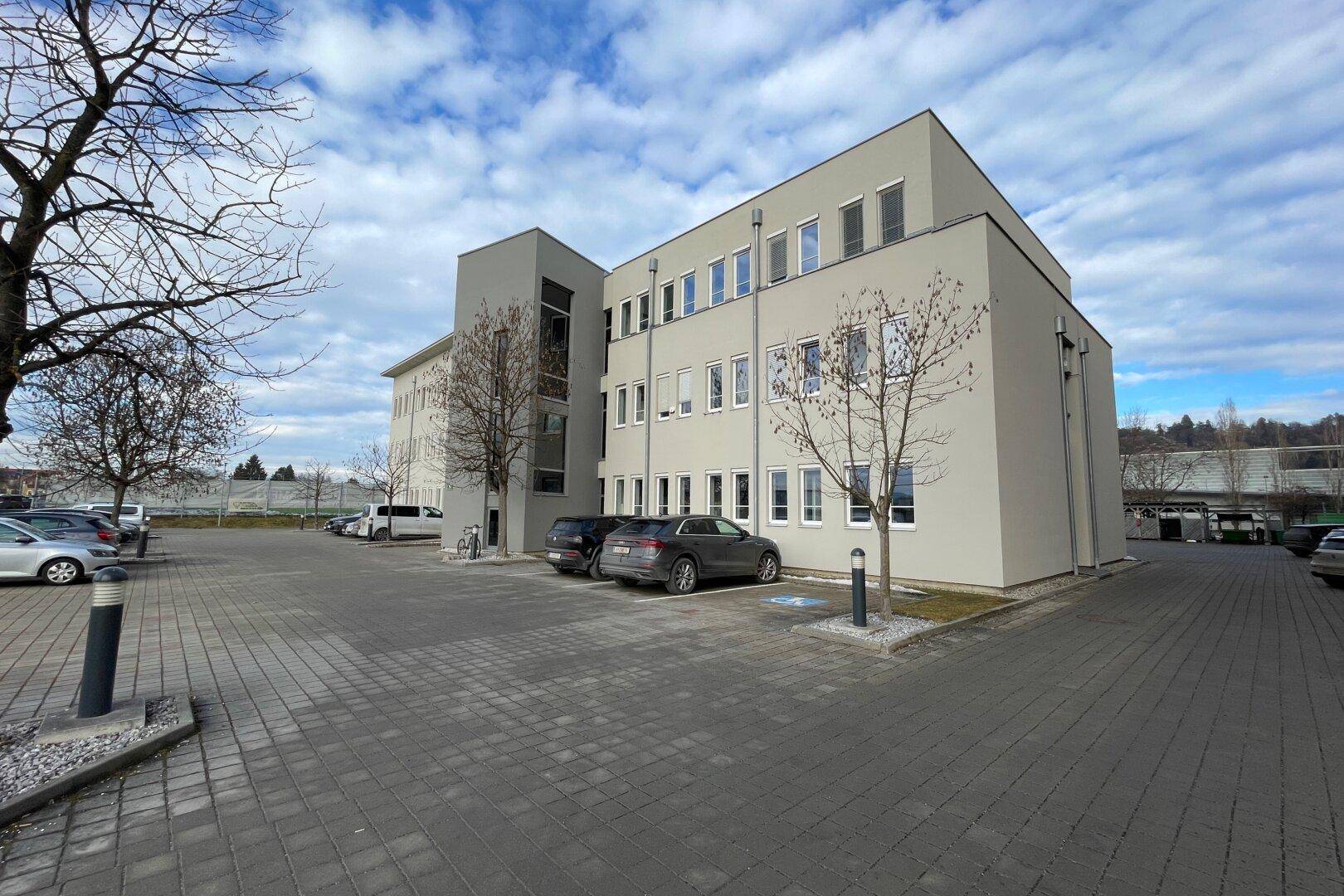Modernes Bürogebäude mit heller Fassade, vielen Fenstern und einem großen Parkplatz davor.