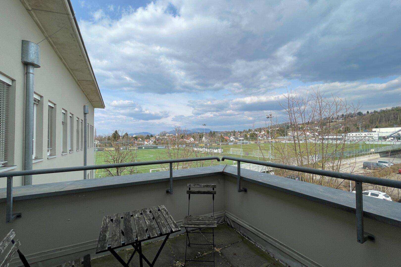 Kleiner Balkon mit Holztisch und Stühlen, bietet Blick auf ein Fußballfeld und die umliegende Landschaft.