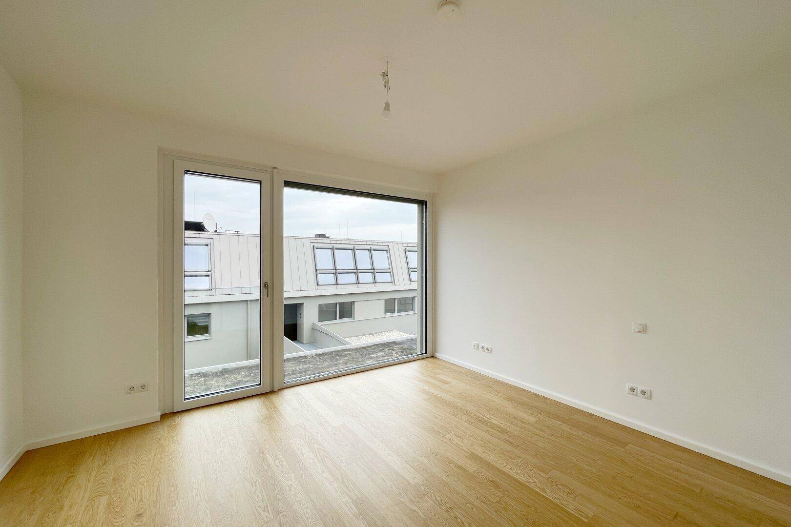 Helles Schlafzimmer mit Parkettboden und großem Fenster mit Blick auf Dächer.