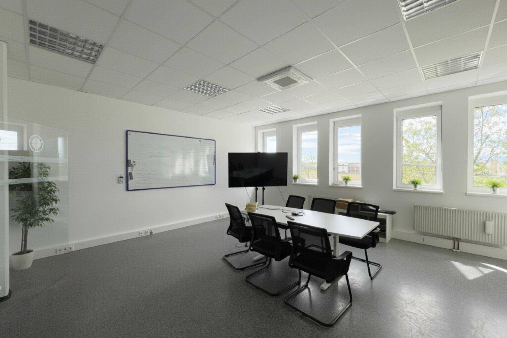 Konferenzraum mit großem Bildschirm, Whiteboard und viel Tageslicht durch die Fenster.