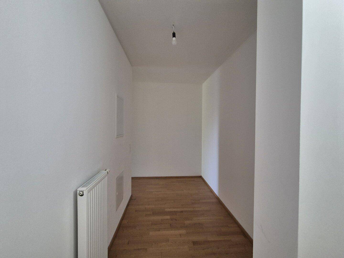 Schmaler Flur mit Holzparkettboden, weiß gestrichenen Wänden und Heizkörper.