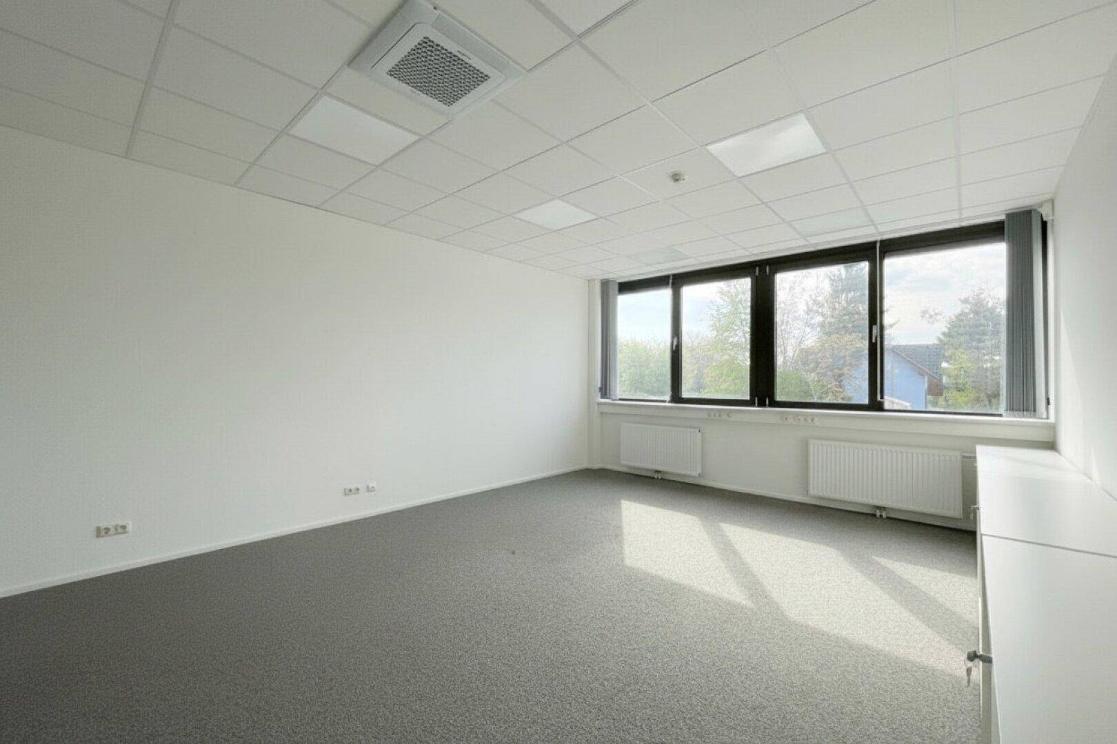 Kleineres Büro mit Teppichboden, drei Fenstern und Klimaanlage an der Decke.