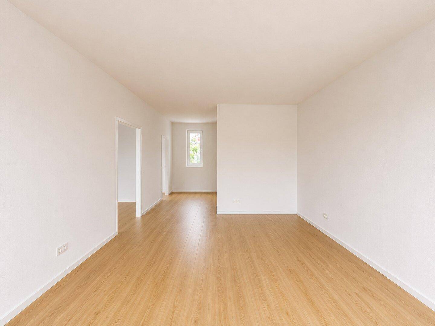 Geräumiges, leeres Zimmer mit Laminatboden und Blick in einen angrenzenden Flur.